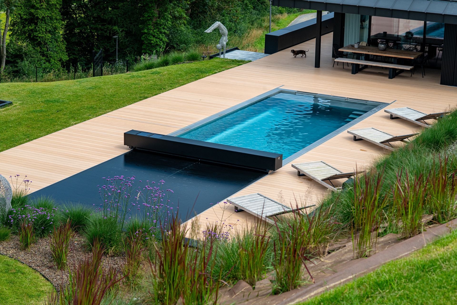 A rectangular swimming pool with a partially extended cover on a wood deck next to a lawn and modern outdoor dining area.