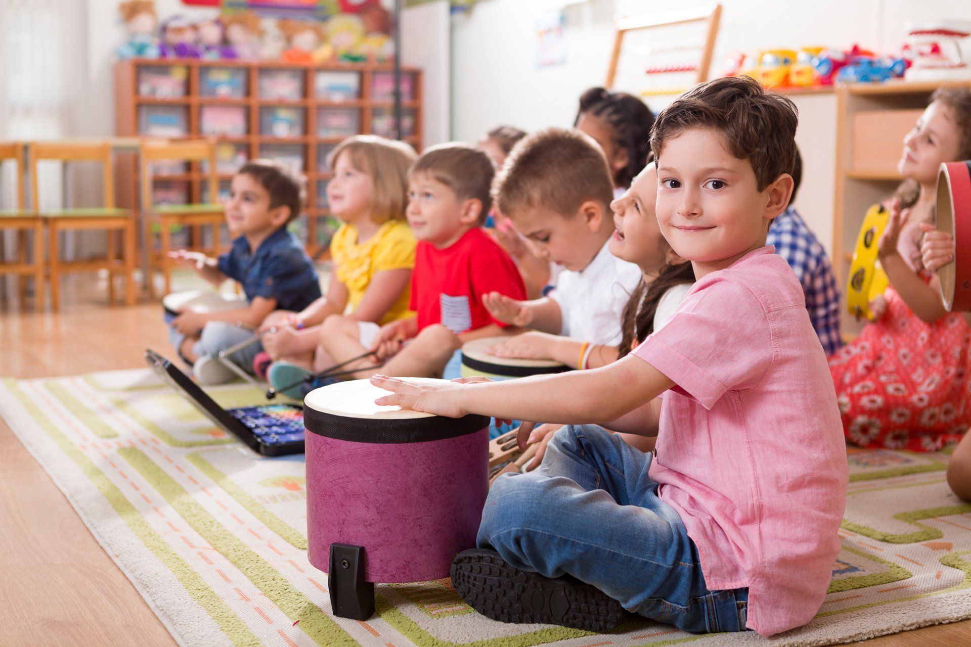 I bambini in classe suonano strumenti: tamburi, tamburello. Un bambino sorride. Tappeto, stanza colorata.