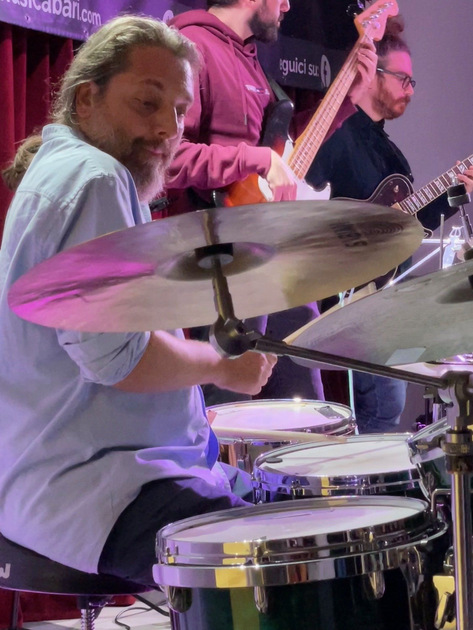 Batterista che suona con una band. Luci viola. Altri membri della band con le chitarre sullo sfondo.