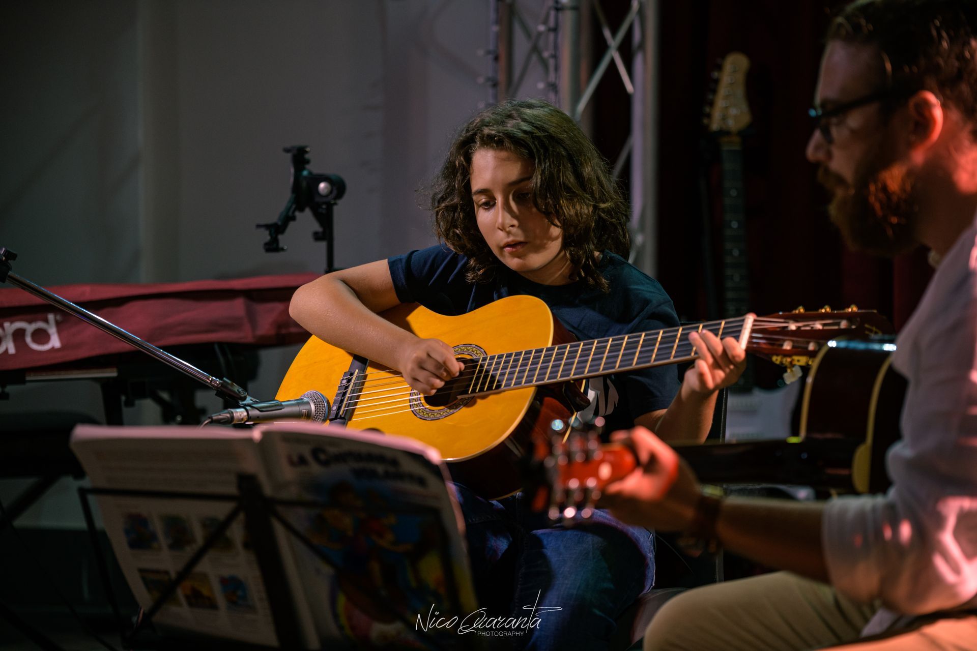 Un ragazzo suona una chitarra acustica gialla con un insegnante. Si trovano in una stanza scarsamente illuminata, con strumenti musicali.