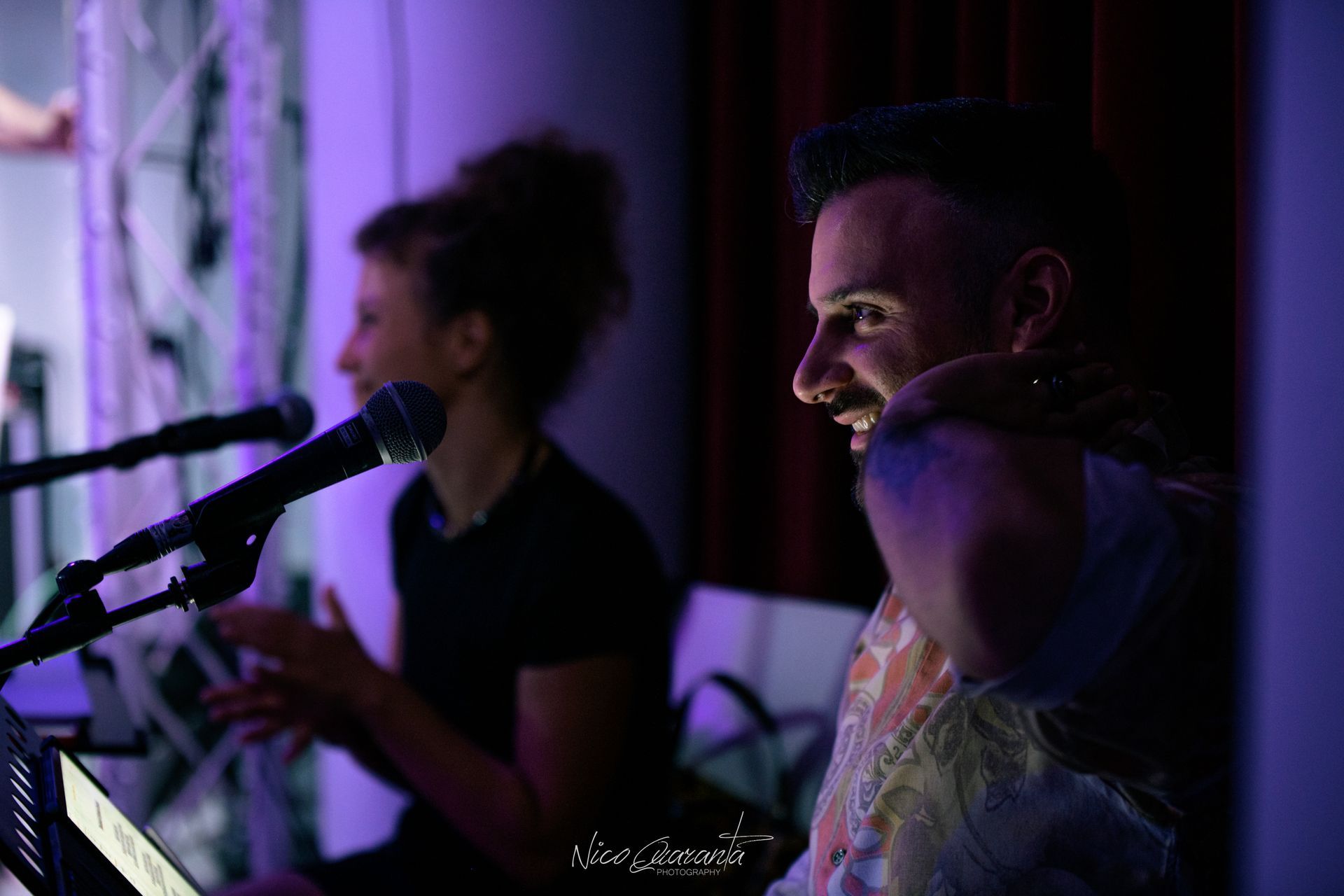 Due persone si esibiscono con i microfoni in una stanza scarsamente illuminata, sorridenti.