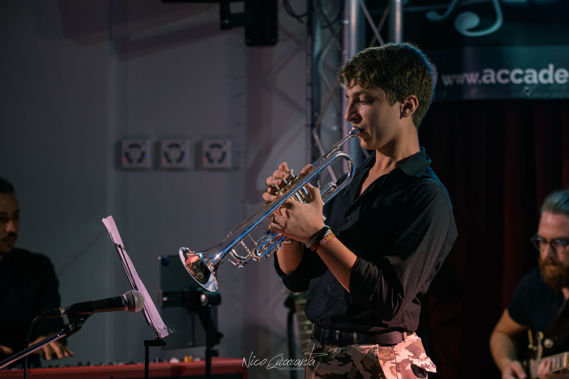 Un musicista suona una tromba d'argento sul palco, con un leggio e i compagni di band visibili.