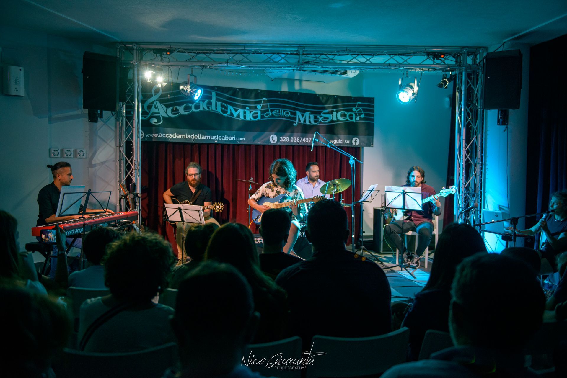 La band suona sul palco con il pubblico seduto di fronte. Luci blu sul palco, striscioni sullo sfondo.