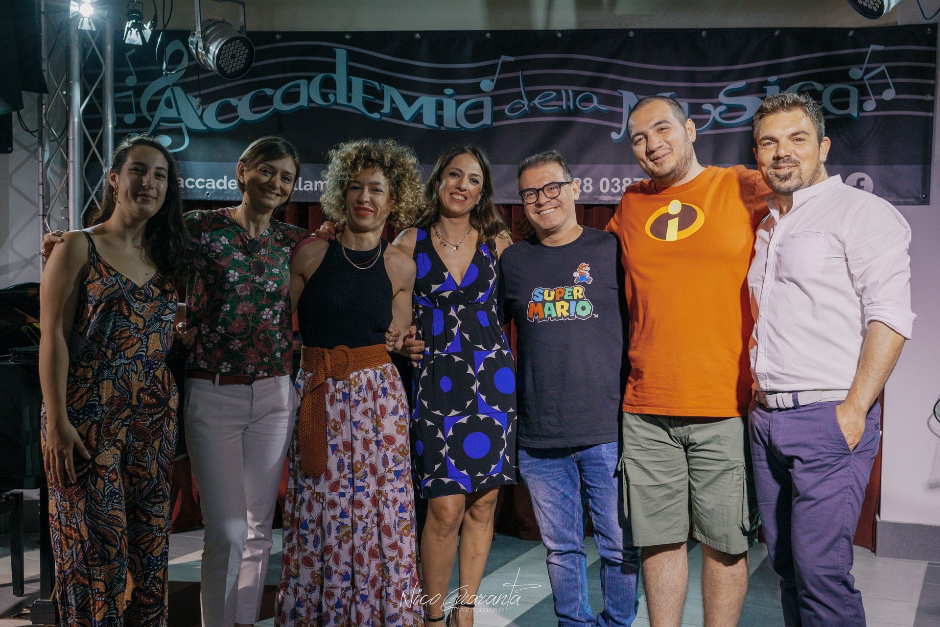 Un gruppo di sette persone sorridenti posa per una foto davanti a uno striscione presso l'accademia musicale.