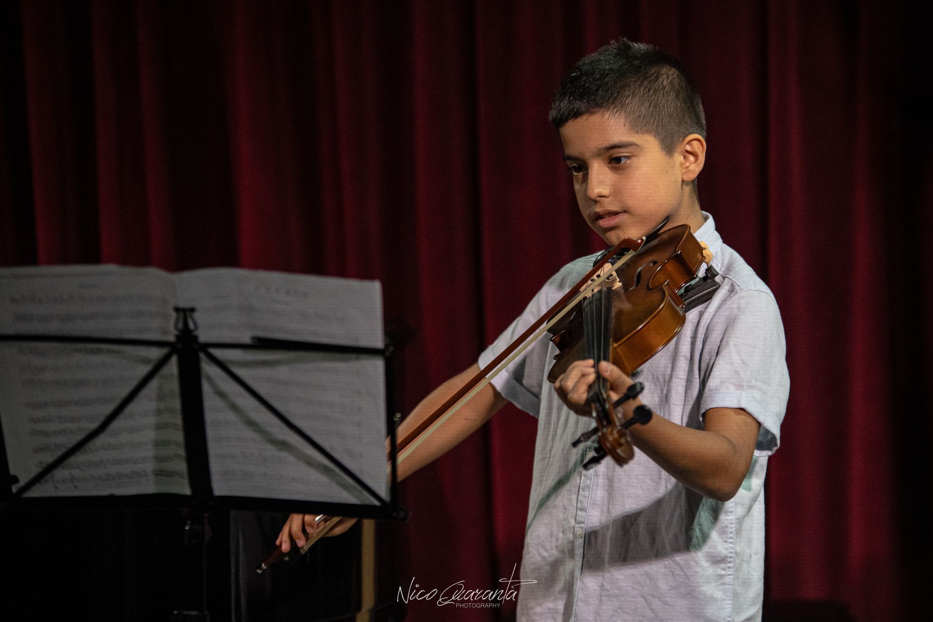 Ragazzo che suona il violino su un palco con spartiti; sullo sfondo una tenda rossa.