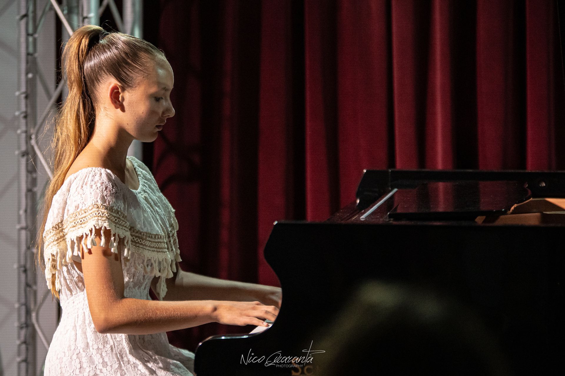 Una donna suona un pianoforte a coda sul palco. Indossa un abito bianco, ha i capelli raccolti in una coda di cavallo e sullo sfondo si vedono delle tende rosse.