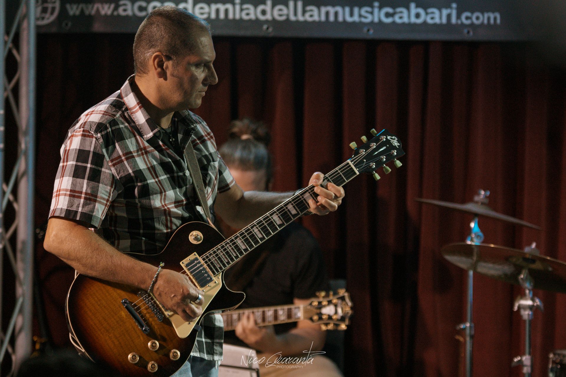 Uomo che suona la chitarra elettrica sul palco; band sullo sfondo; ambiente buio.