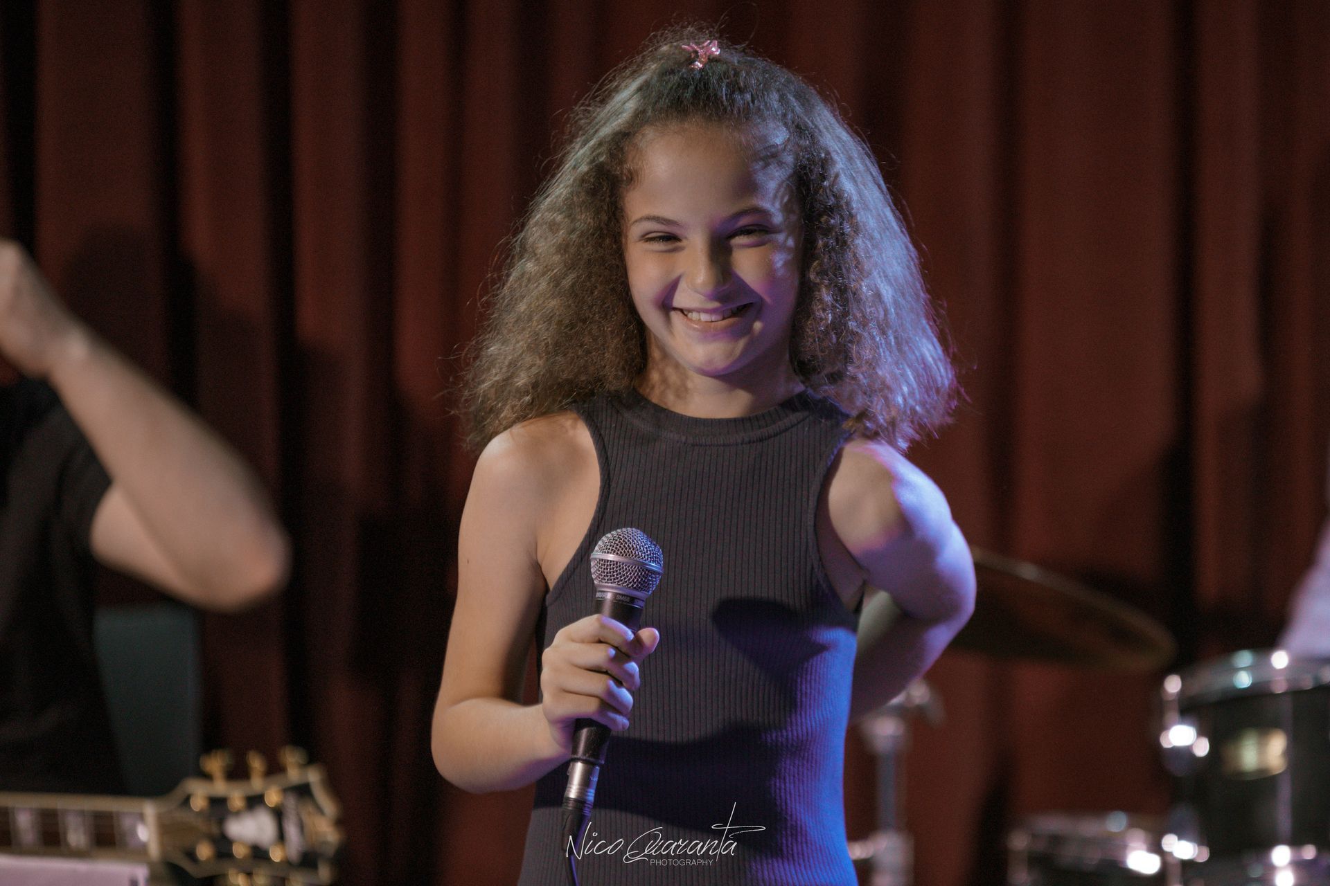 Una ragazza sorridente canta al microfono sul palco, con una band e una tenda rossa sullo sfondo.