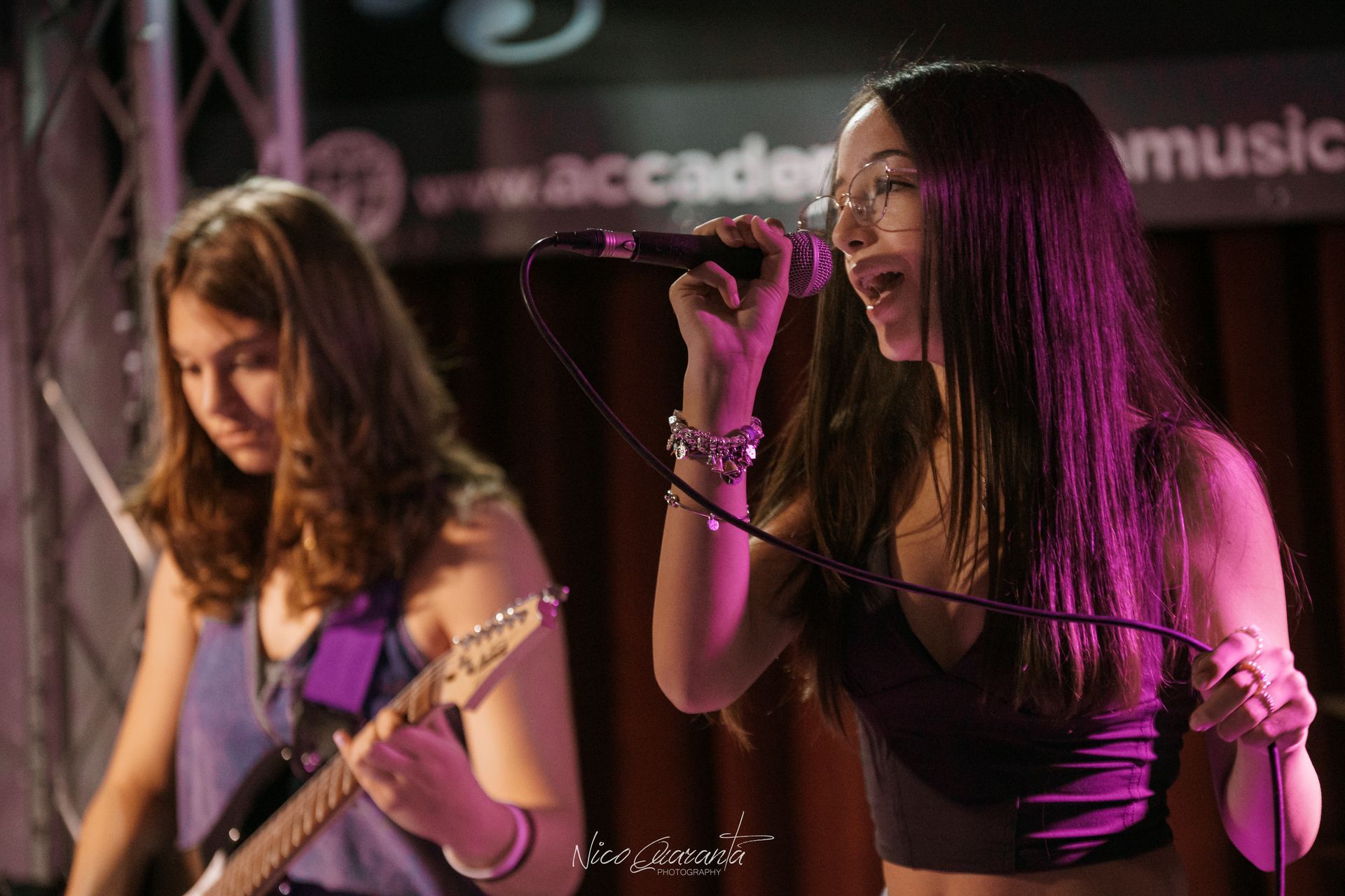 Una donna canta al microfono; un'altra suona la chitarra sul palco, sotto una luce viola.