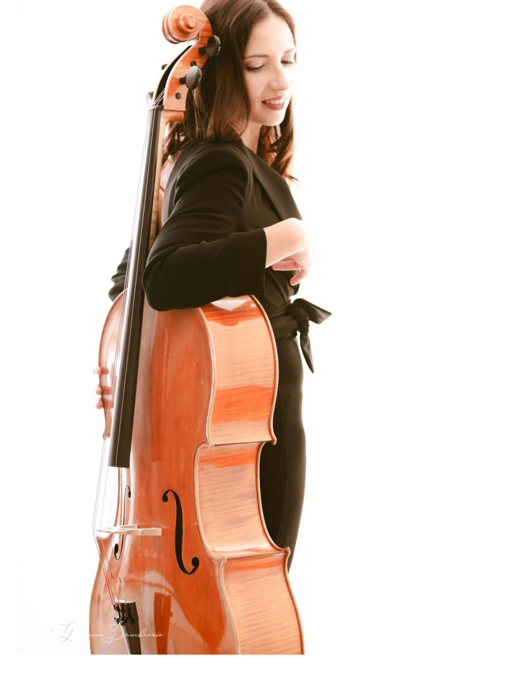 Donna vestita di nero appoggiata a un violoncello. Sorridente, con lo strumento su sfondo bianco.