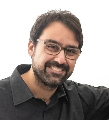 Uomo con occhiali e barba, sorridente. Indossa una camicia nera con colletto, su sfondo bianco.