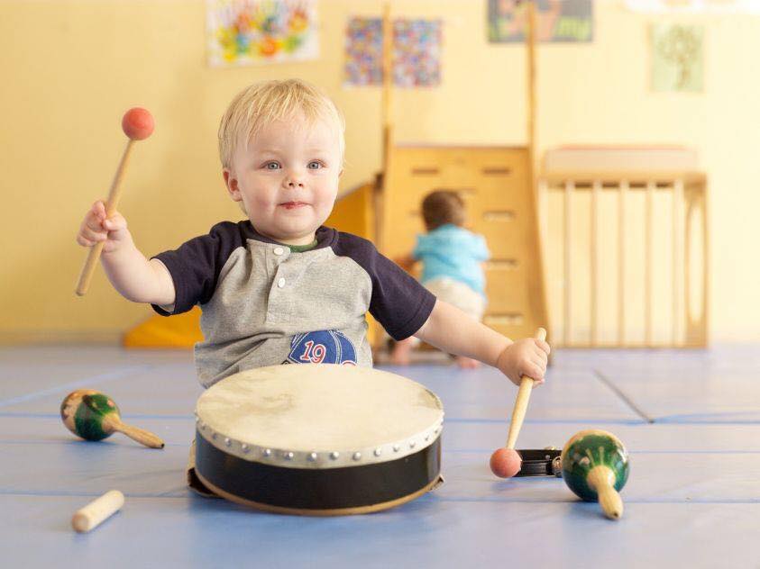 Un bambino piccolo suona felicemente un tamburo, tenendo in mano le bacchette, con delle maracas nelle vicinanze. Gioco al chiuso.