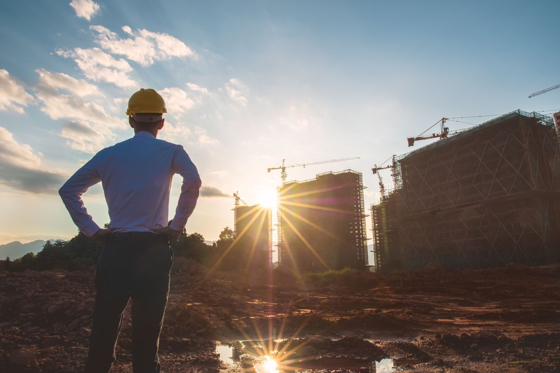 Construction worker in hard hat stands at a building site at sunset, facing cranes and scaffolding.