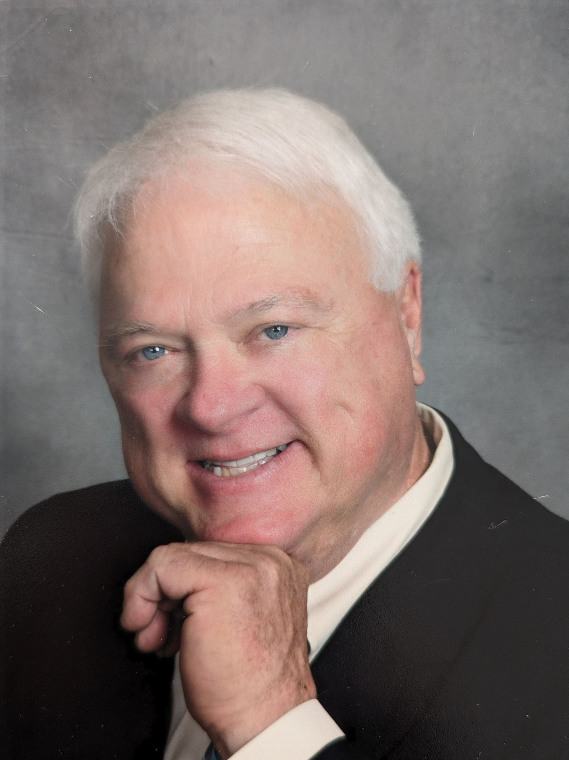 Smiling person with white hair resting chin on hand against a gray background