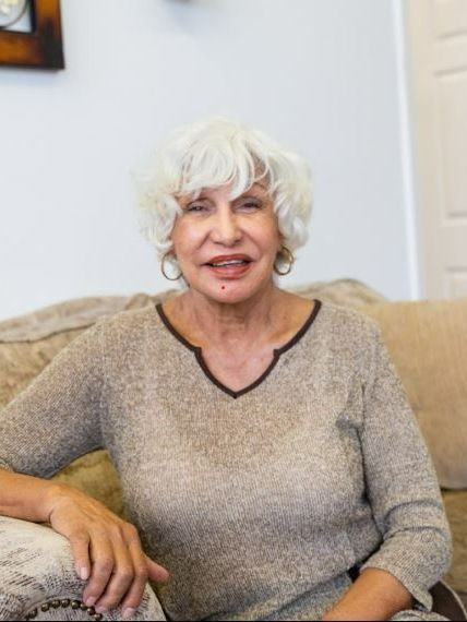Smiling person with white hair sitting on a beige couch in a light-colored room