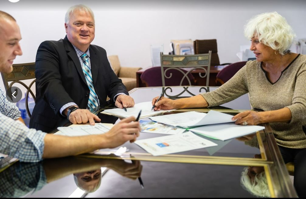 Three people in a meeting reviewing papers around a glass table, one writing with a pen