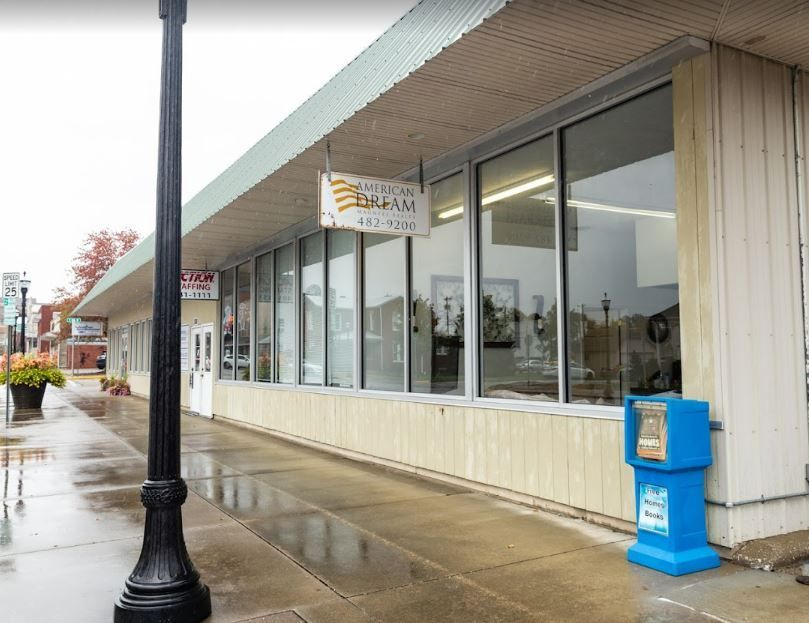 Rainy sidewalk outside a strip mall with glass storefronts, a lamppost, and a blue newspaper box