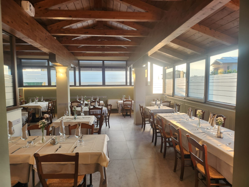Luminosa sala da pranzo del ristorante con travi in legno, lunghi tavoli apparecchiati e luce naturale che filtra dalle grandi finestre.