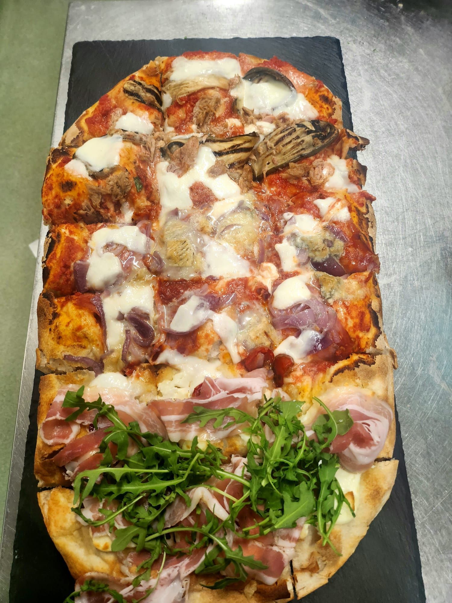 Pizza rettangolare assortita su un tagliere scuro con formaggio, pancetta, verdure e olive.
