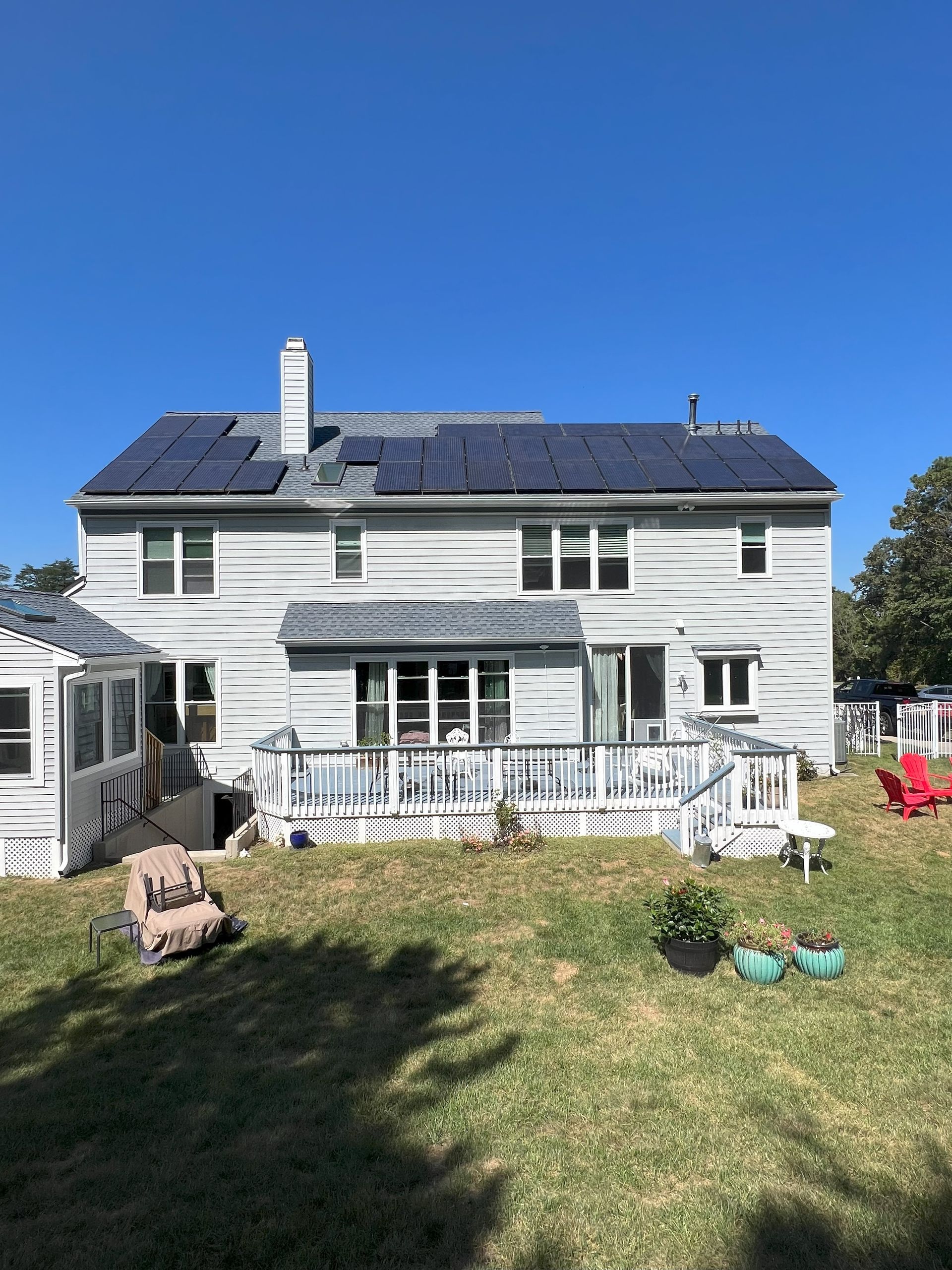 White two-story house with dark roof, white deck, and grassy yard under a clear blue sky