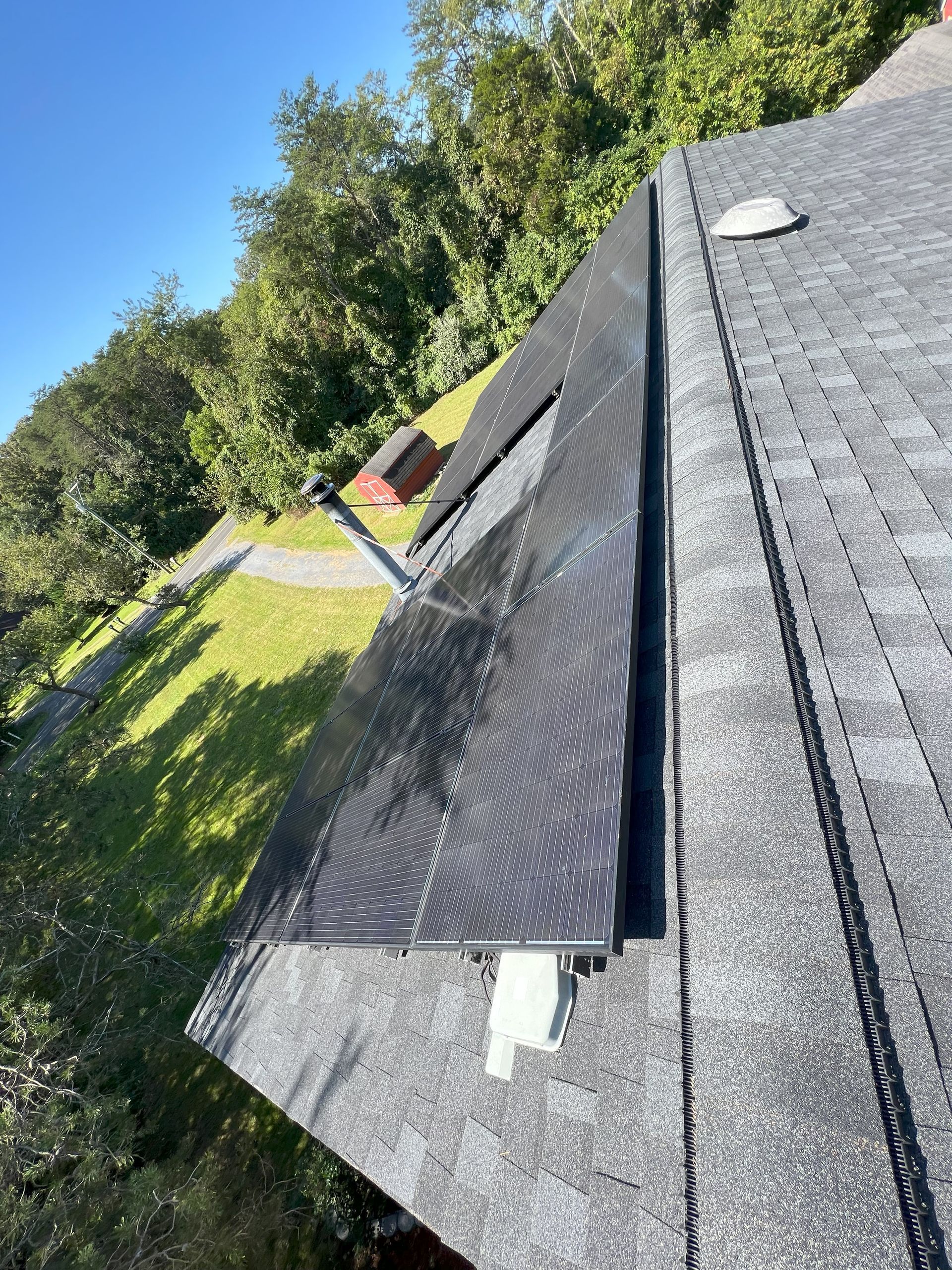 Solar roof panel on a dark shingled rooftop beside trees and lawn under a clear blue sky