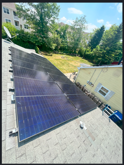 Solar panels installed on a shingled roof beside a house and trees on a sunny day