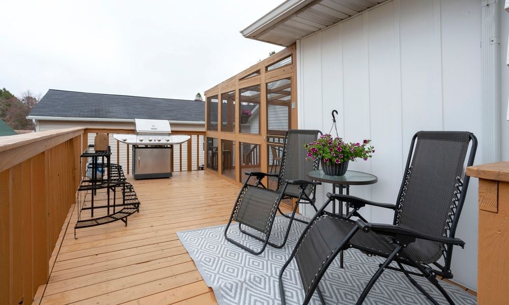 Wooden rooftop deck with black patio chairs, table, grill, and potted flowers beside a white house