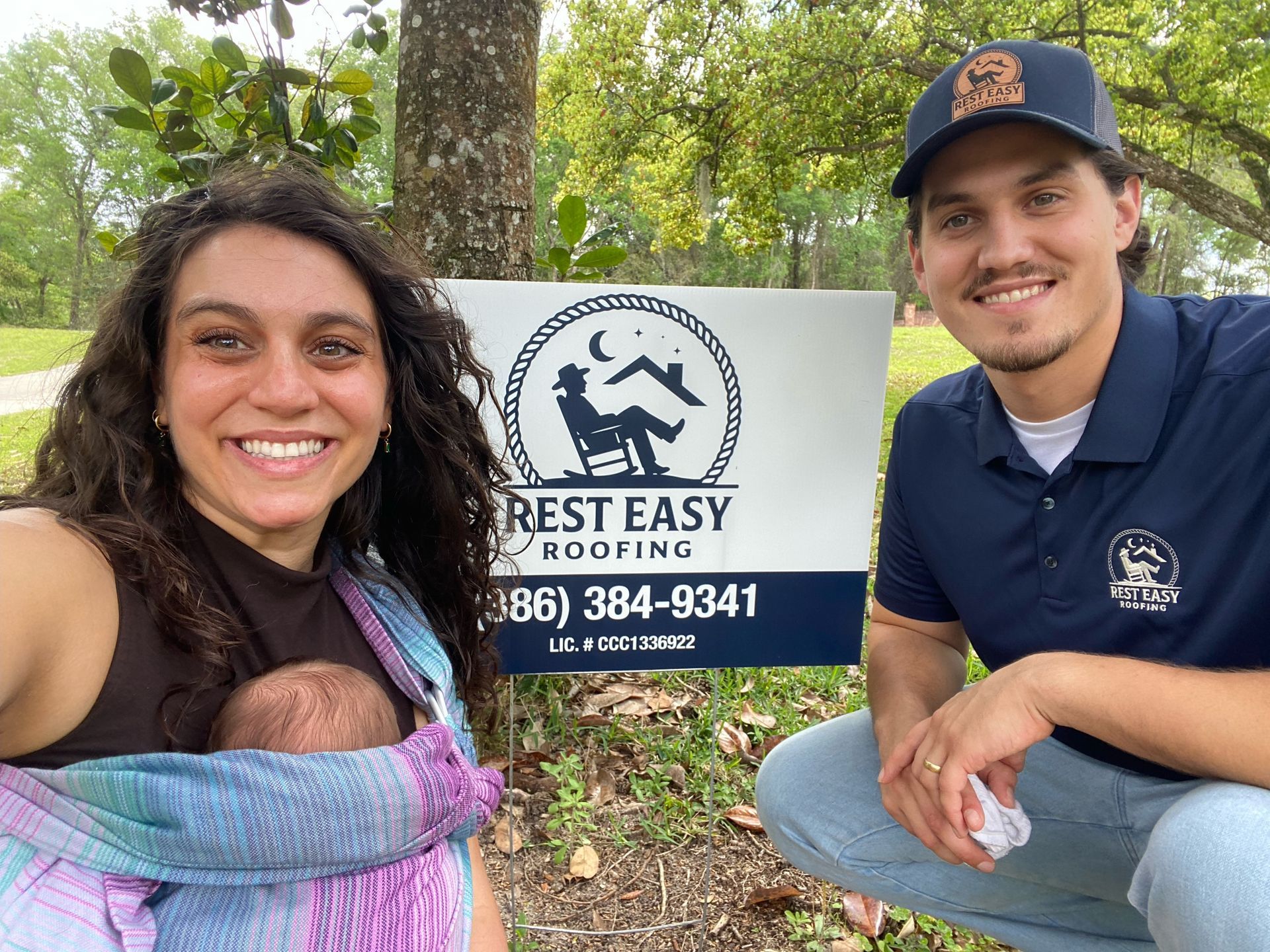 Two people kneel beside a Rest Easy Roofing sign in a wooded area, smiling at the camera.