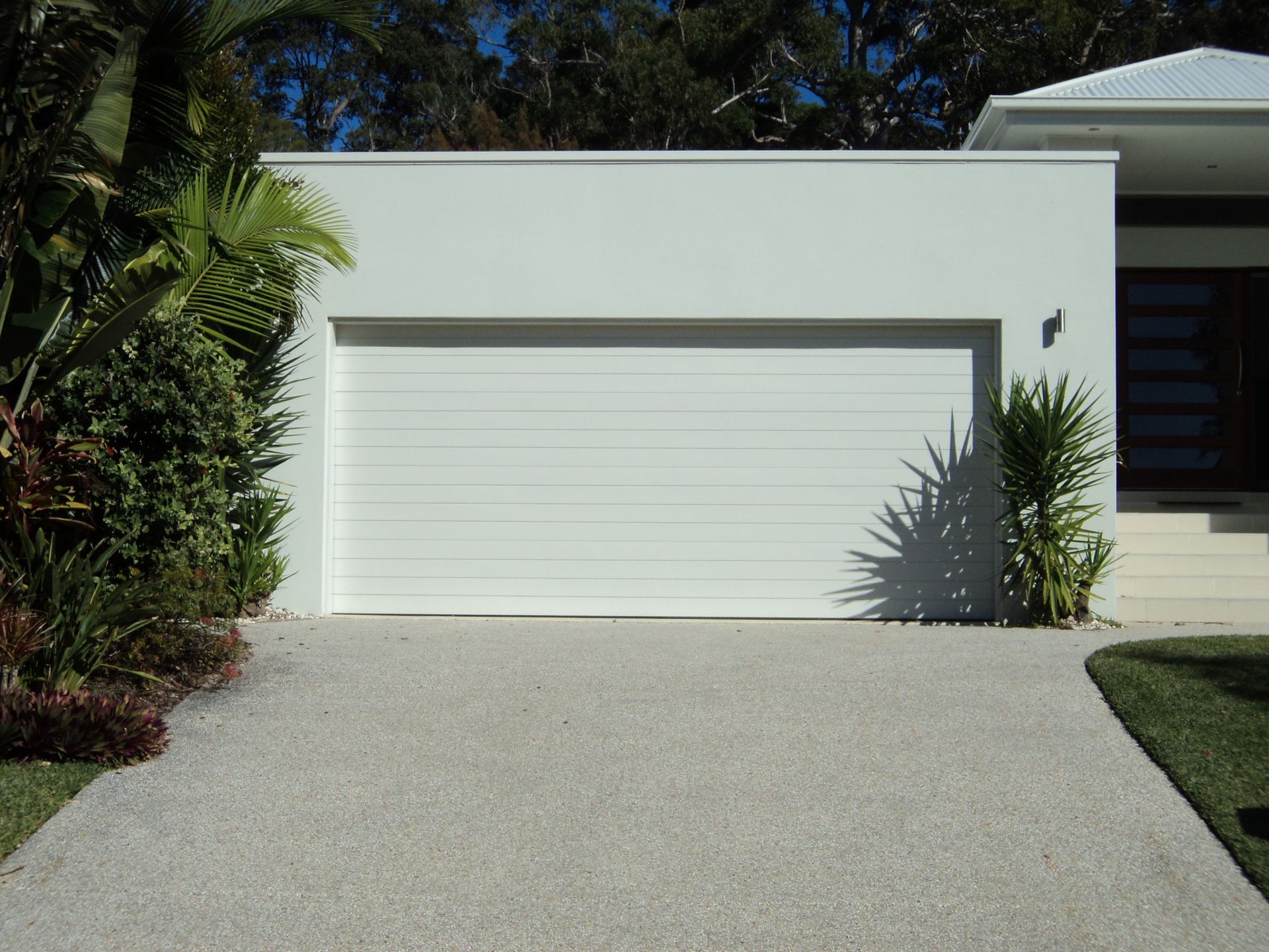 White garage door on a white stucco building with a concrete driveway and landscaping — Coastal Auto Doors & Gates In Buderim, QLD