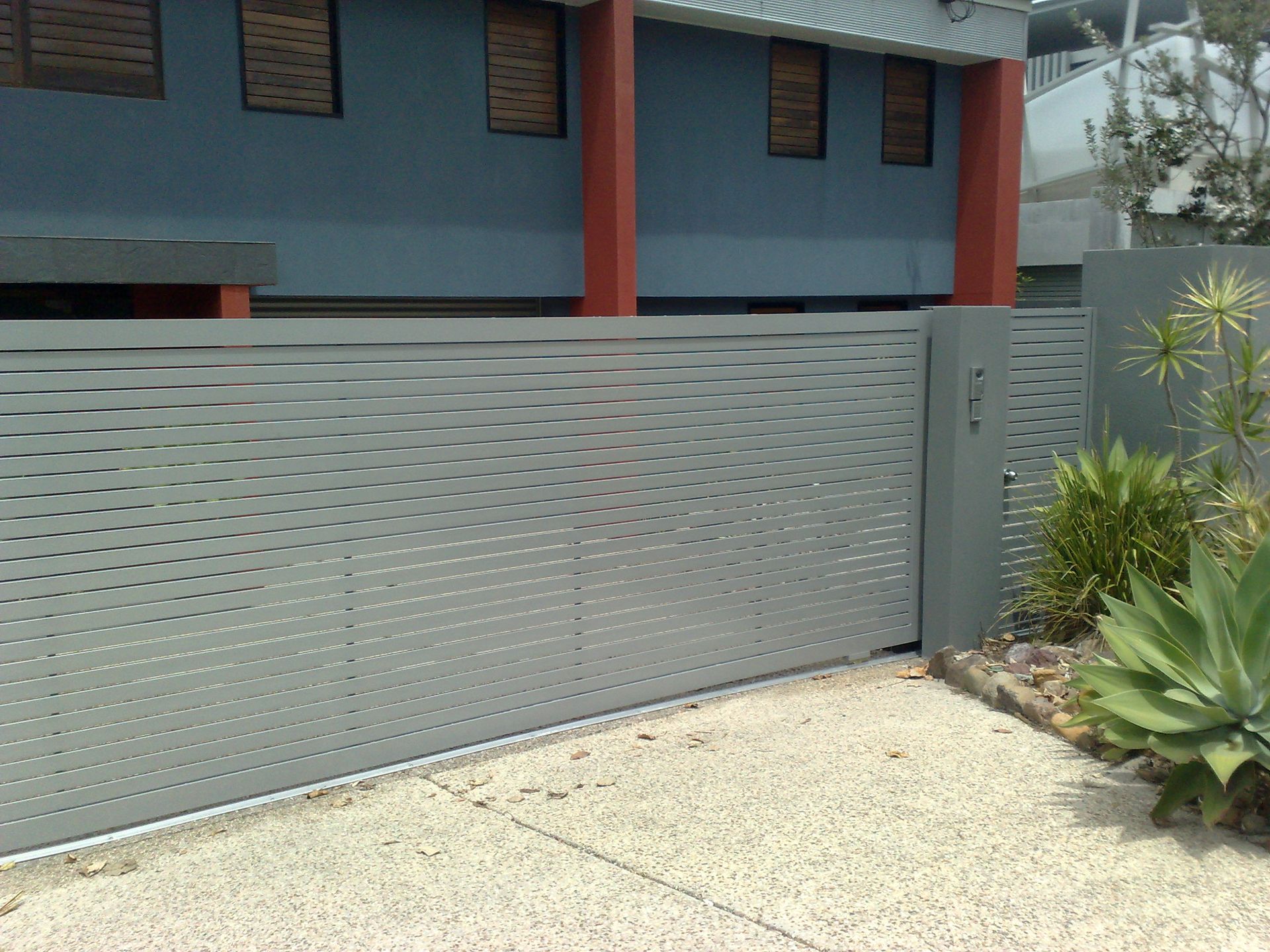 Gray metal sliding gate in front of a blue building with red pillars — Coastal Auto Doors & Gates In Nambour, QLD