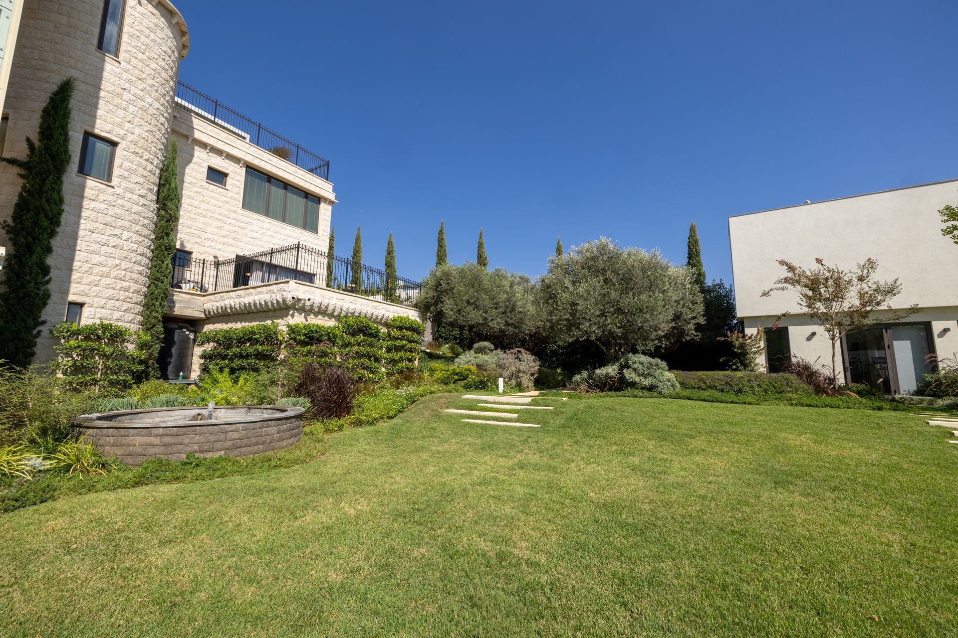 Lush green lawn with stone patio, trees, and buildings against a clear blue sky.