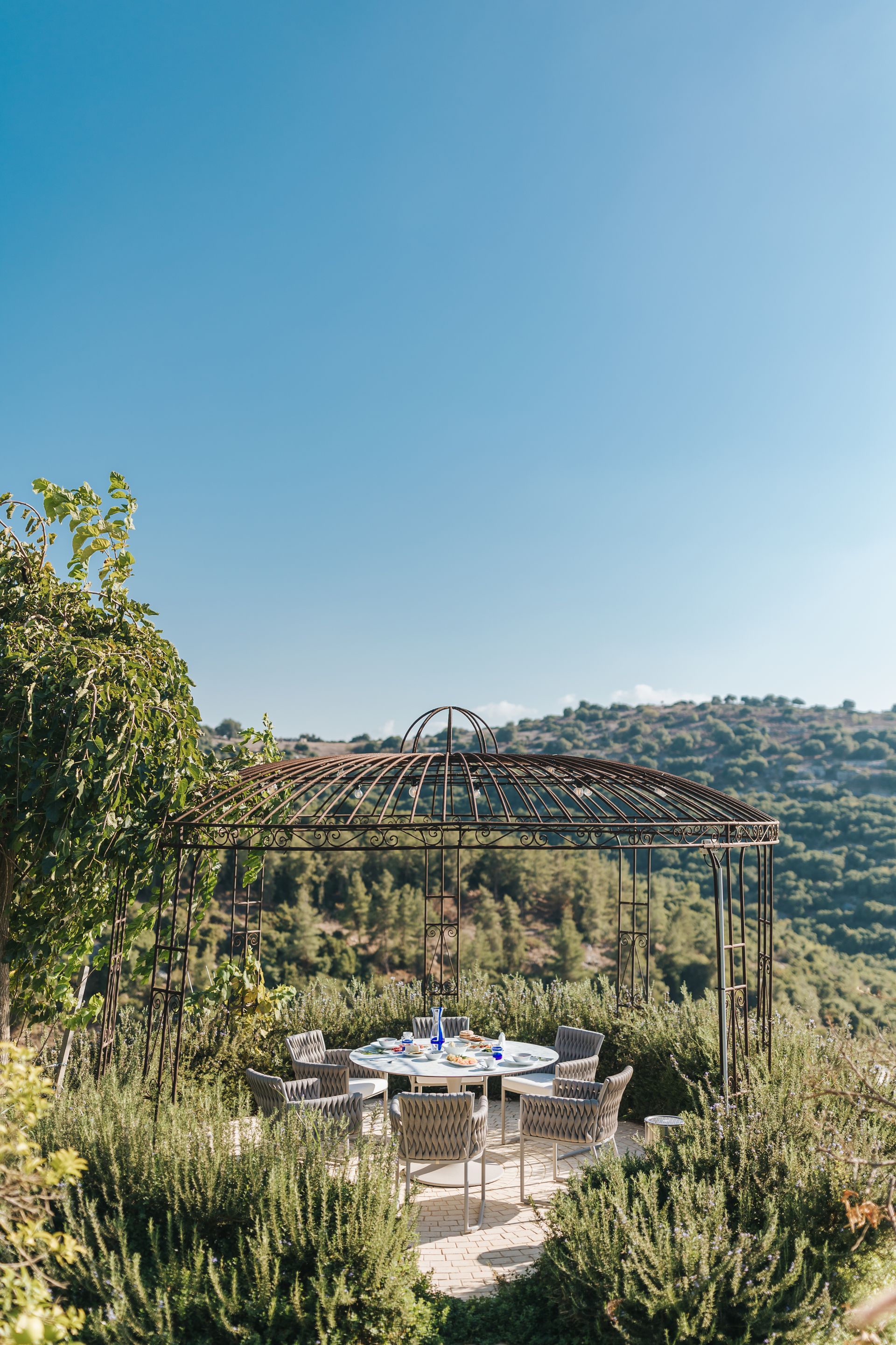 Outdoor dining table under a pergola with blue sky and hillside view.