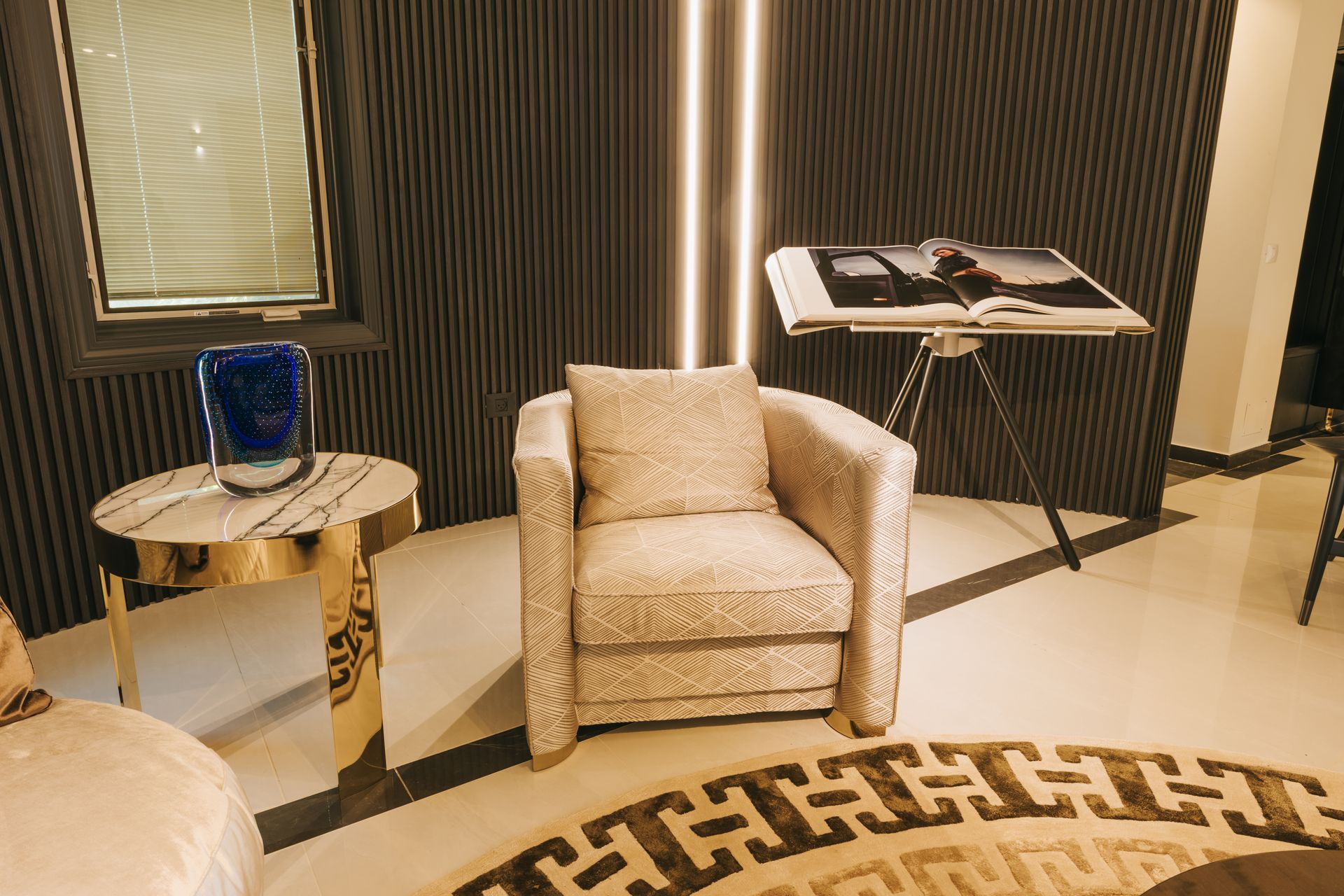 Elegant living room: cream armchair, patterned rug, gold side table with blue vase, and art display.