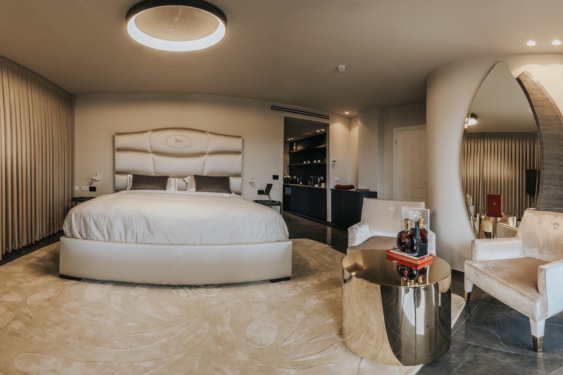 Luxurious bedroom with king-sized bed, seating area, and round gold table. Soft neutral tones and warm lighting.