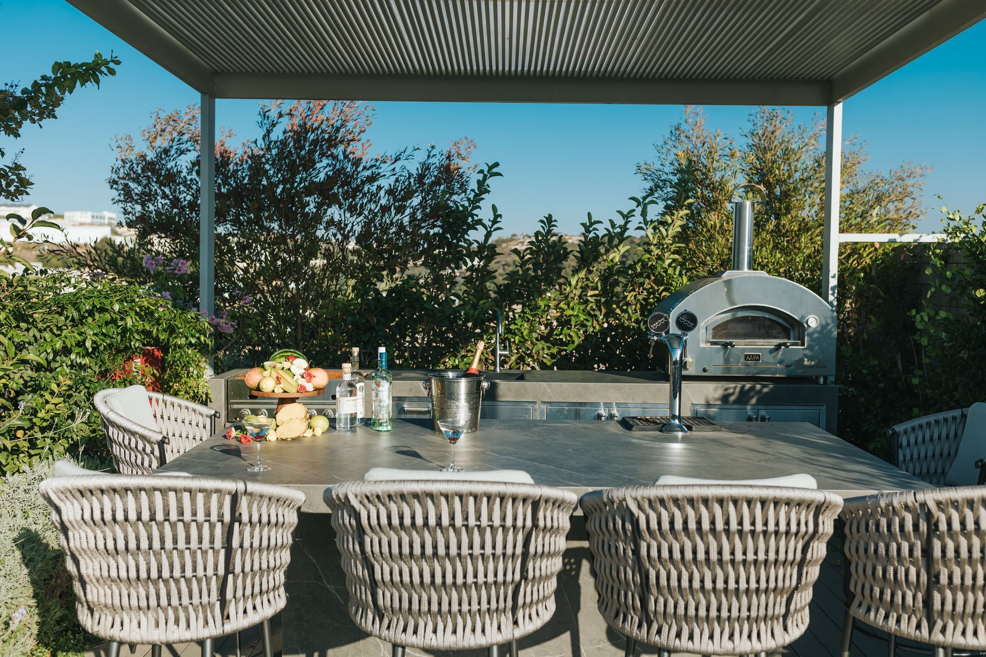 Outdoor kitchen with pizza oven, built-in counter with bar stools, and a pergola overhead.