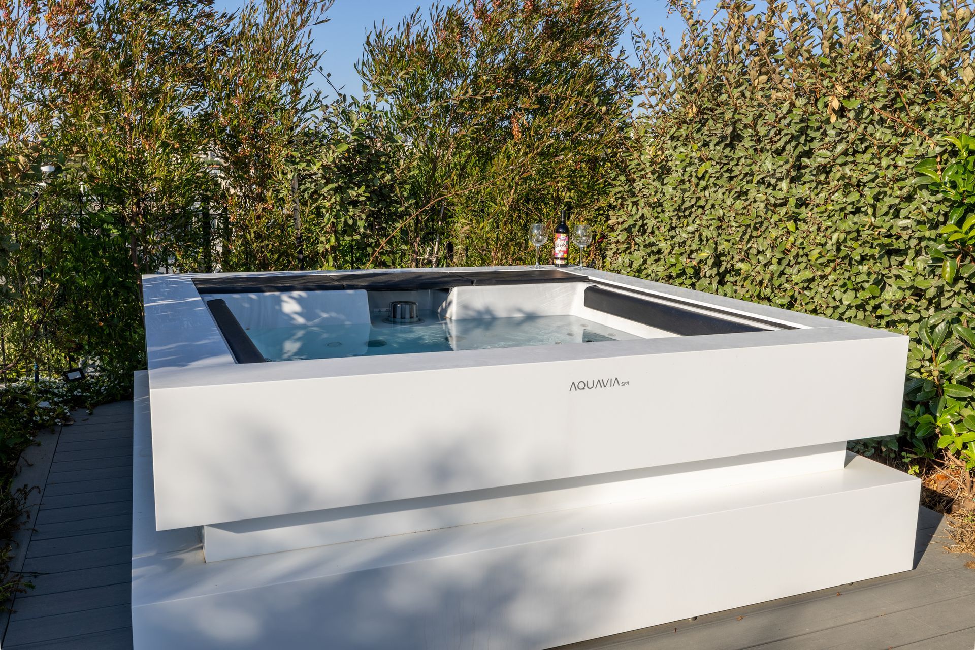 White rectangular hot tub filled with water, outdoors with green foliage in the background.