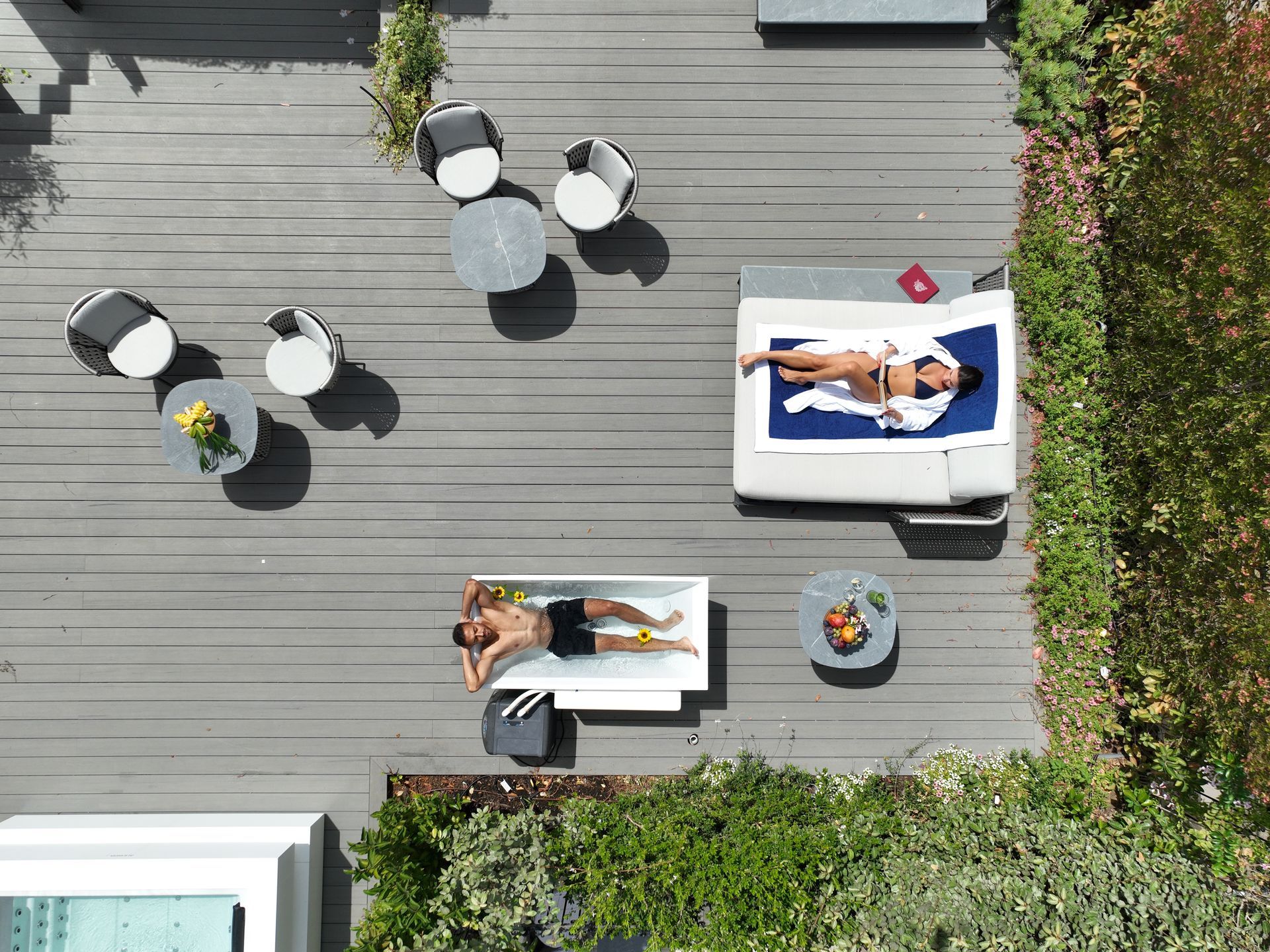 Overhead view of a deck with people sunbathing on lounge furniture near small tables and greenery.
