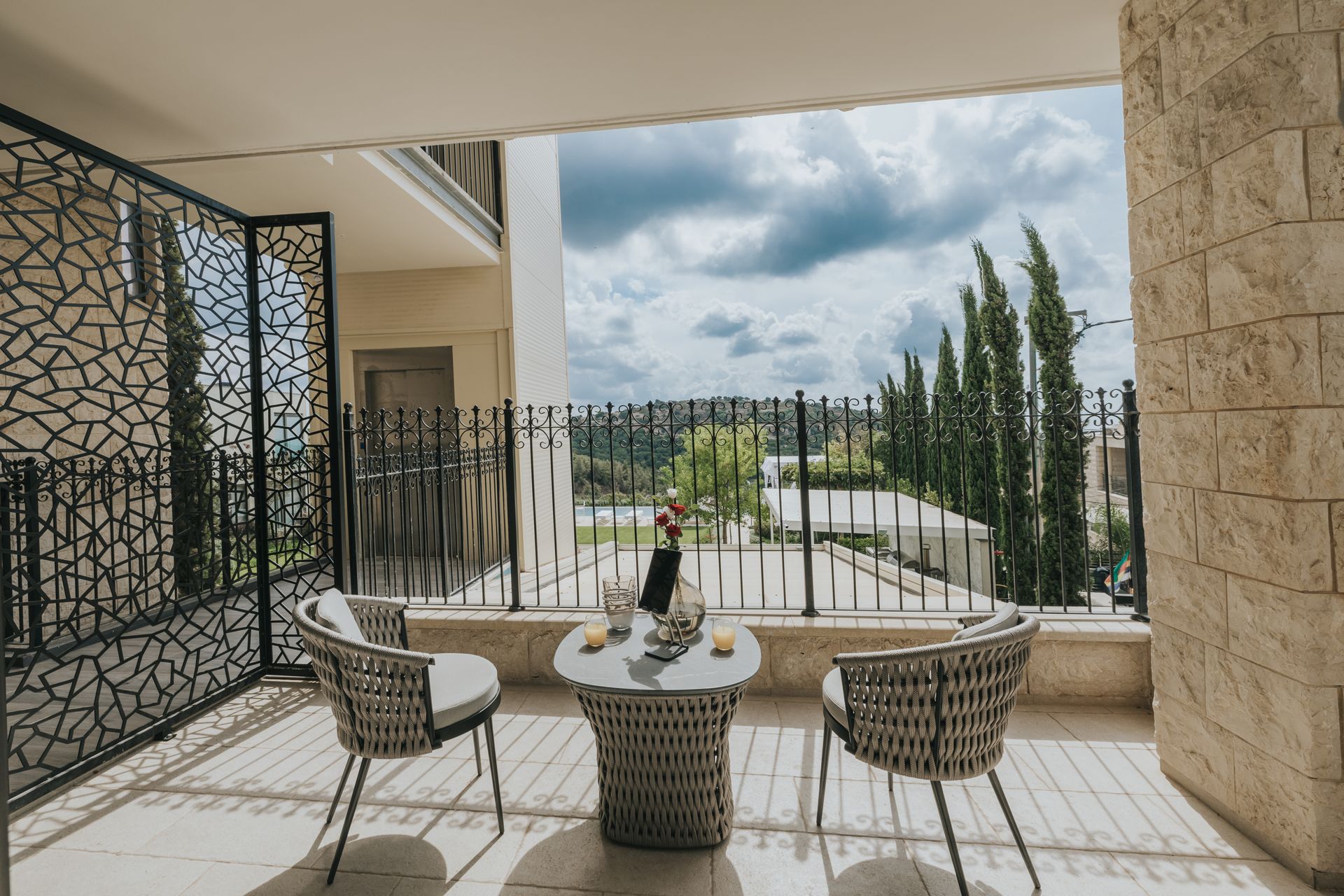Balcony with chairs, small table with drinks, overlooking green landscape with trees and cloudy sky.