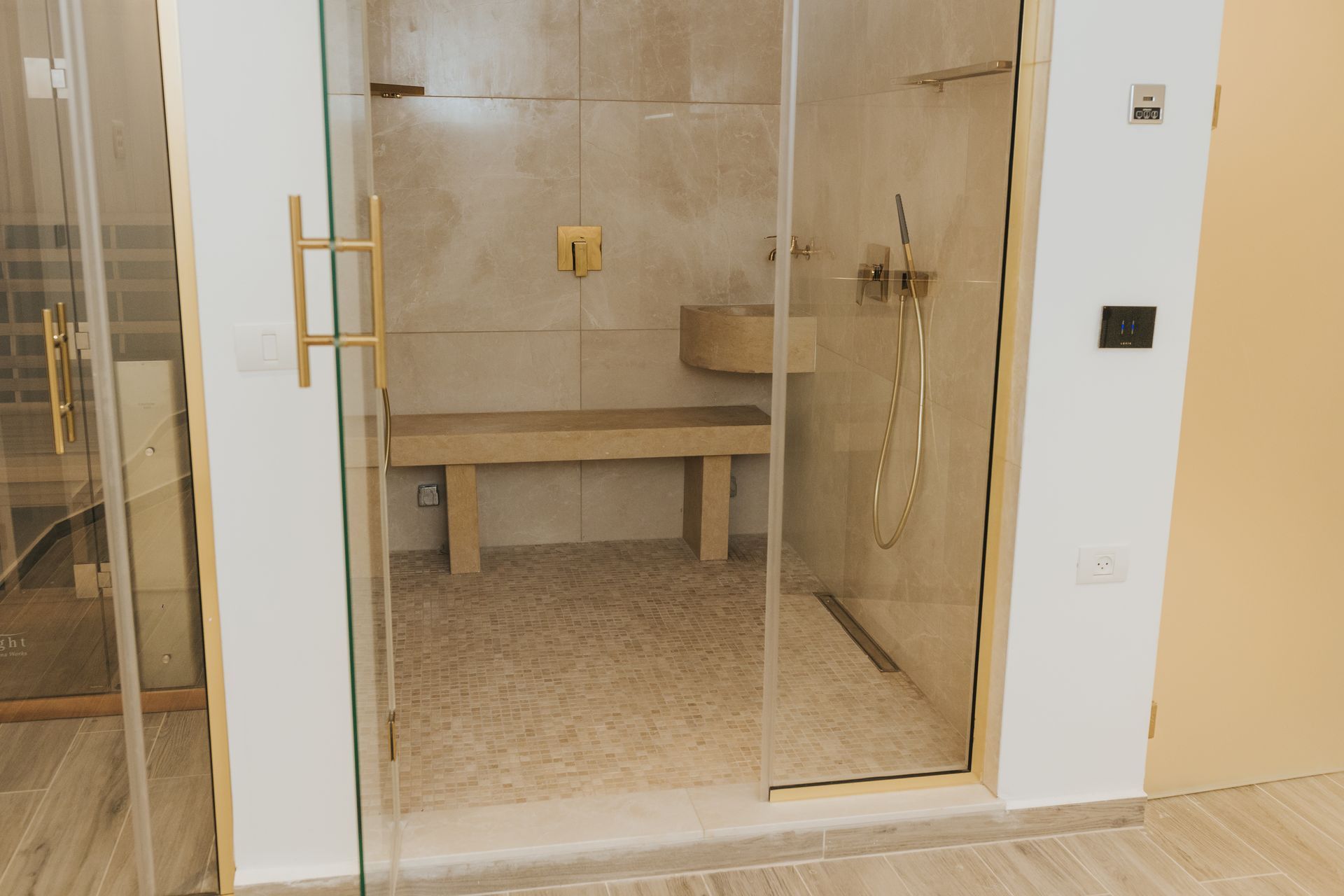 Modern shower with glass doors, stone bench, and gold fixtures. The floor has a light stone texture.