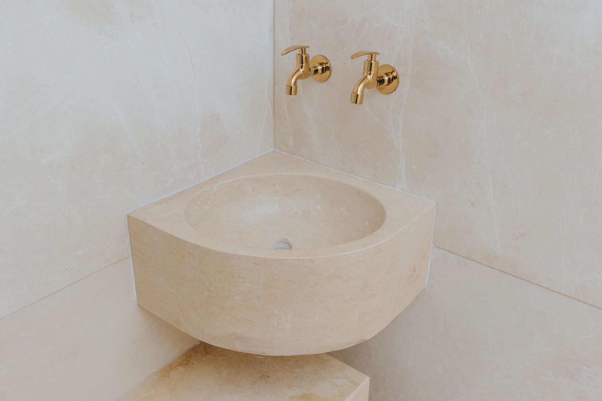Corner stone sink with gold faucets against a light-colored wall.
