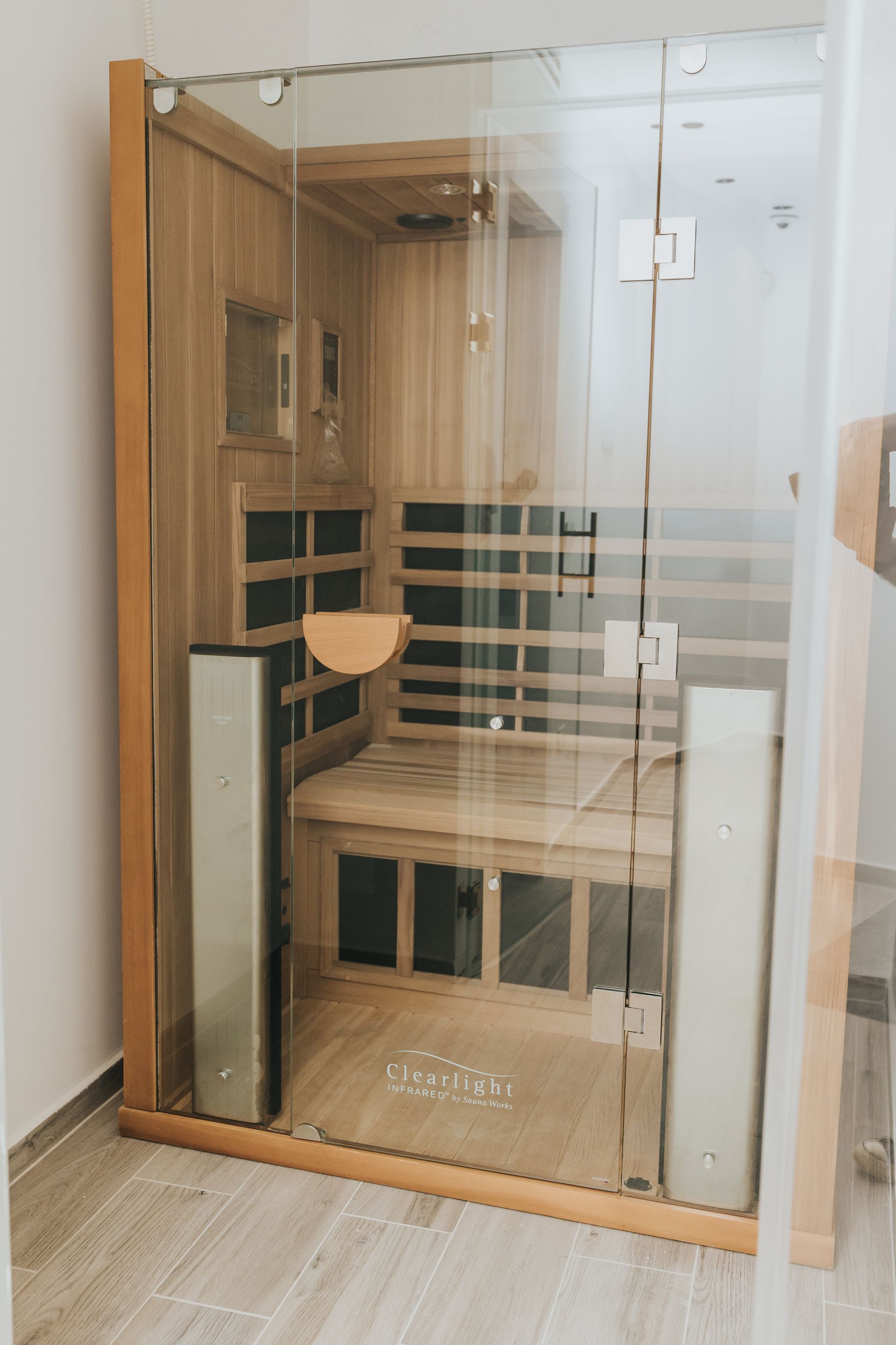 Wooden-framed sauna with glass door, interior wooden benches, and two silver heaters, set on a light-colored floor.
