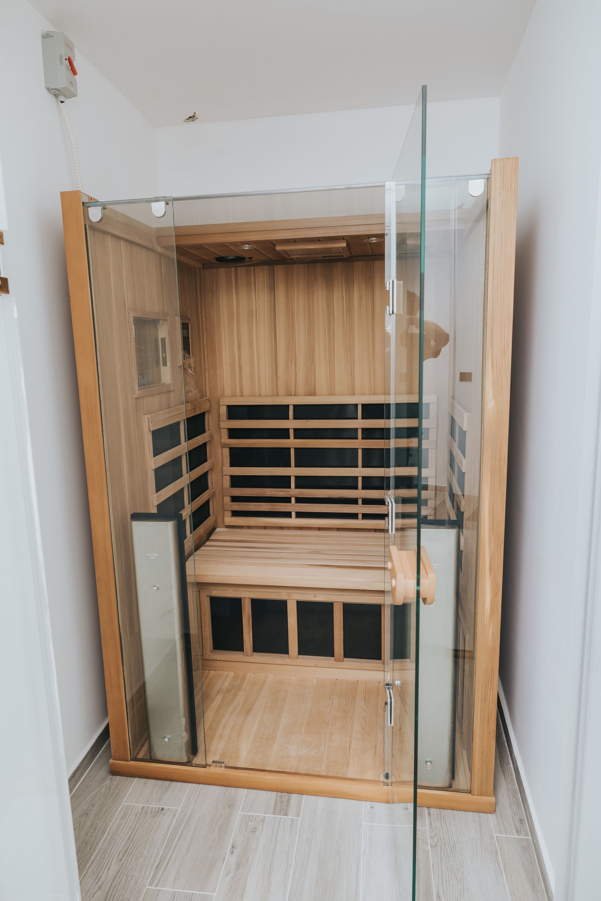 Wooden sauna with glass door in a white-walled room; empty interior with bench seating.