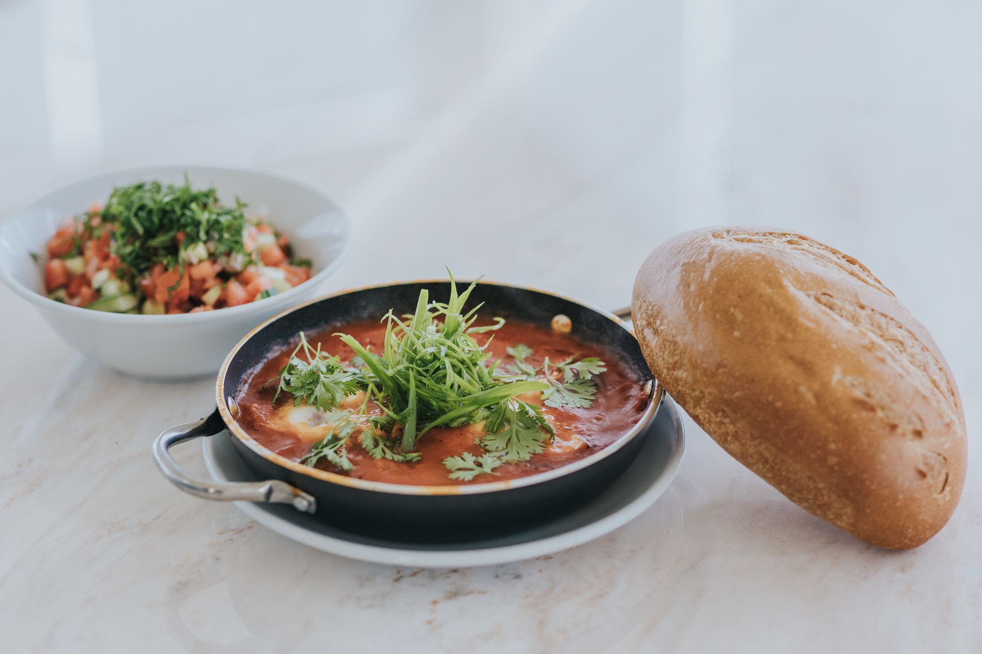 Shakshuka with bread and salad on a white table. Red sauce, green herbs, and a fresh loaf.