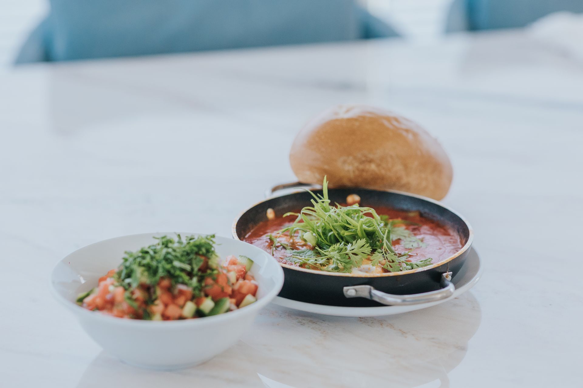 Dish of shakshuka and salad, with bread roll on a white marble surface.