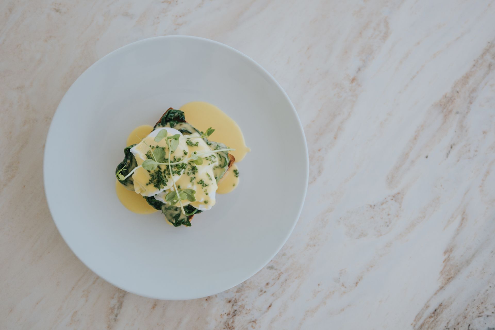 Eggs Benedict on a white plate, with yellow sauce and green spinach, on a marble table.
