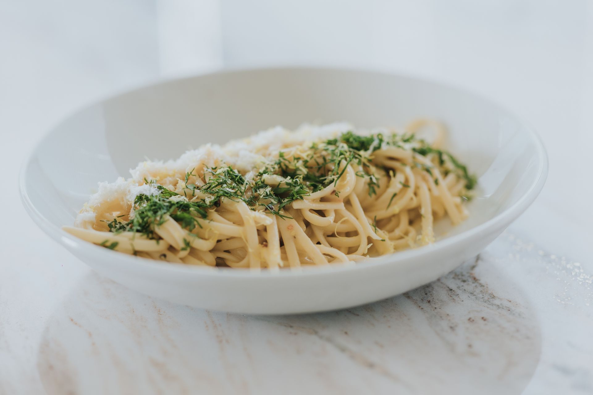Spaghetti in a white bowl, topped with grated cheese and green herbs, on a marble surface.
