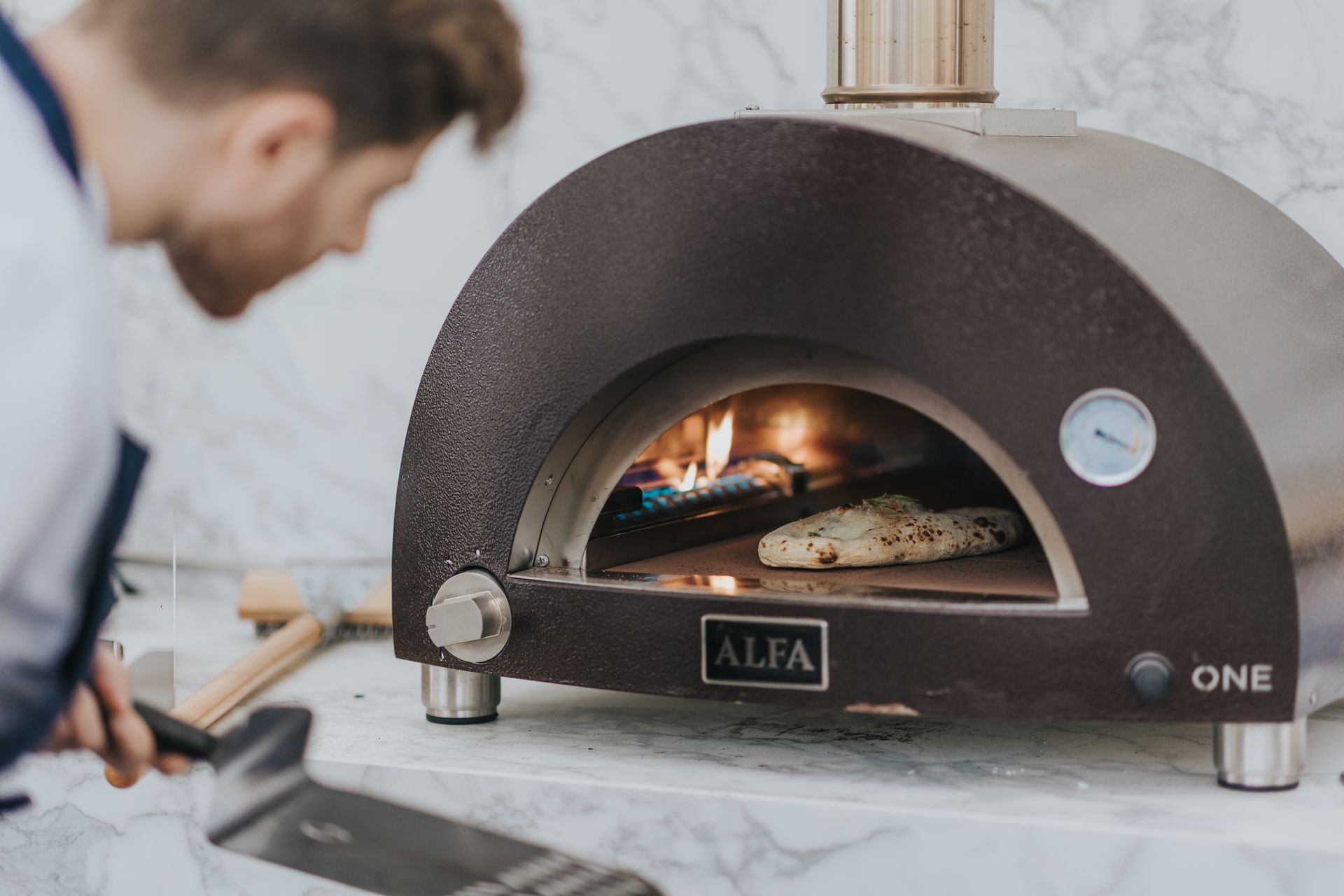 A person using a pizza oven, placing pizza with a peel, flames visible inside.