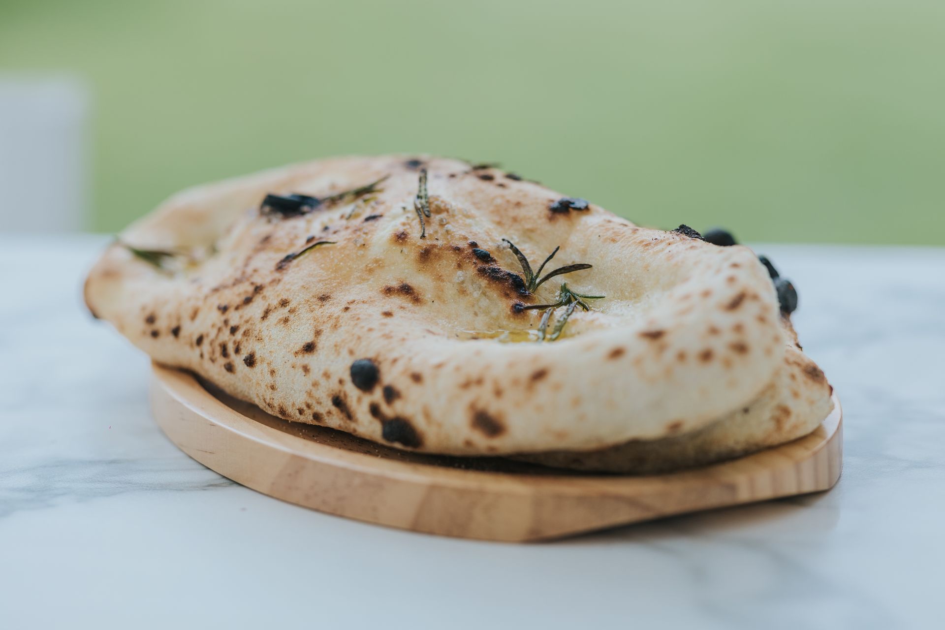 Focaccia bread with herbs and olives on a wooden board against a blurry green background.