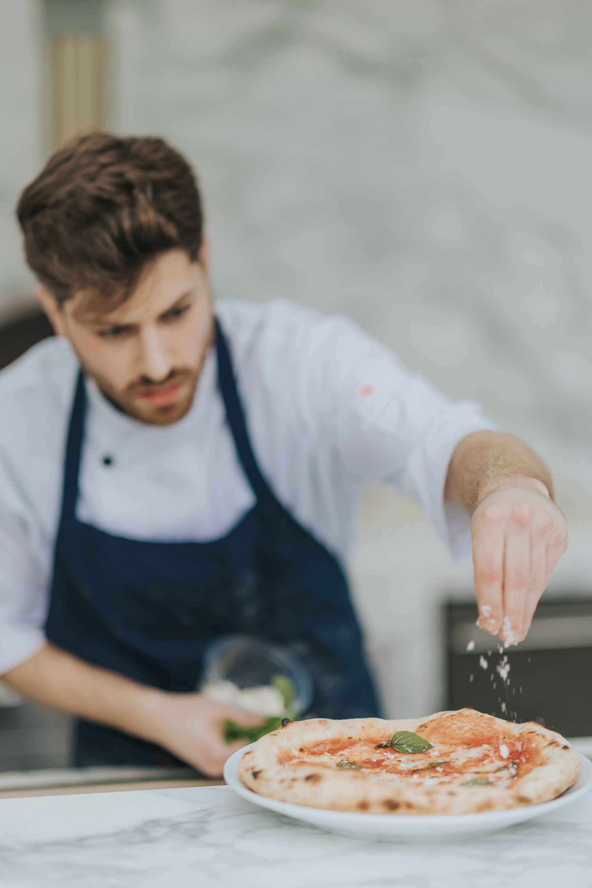 Chef sprinkling cheese on a pizza, kitchen setting.