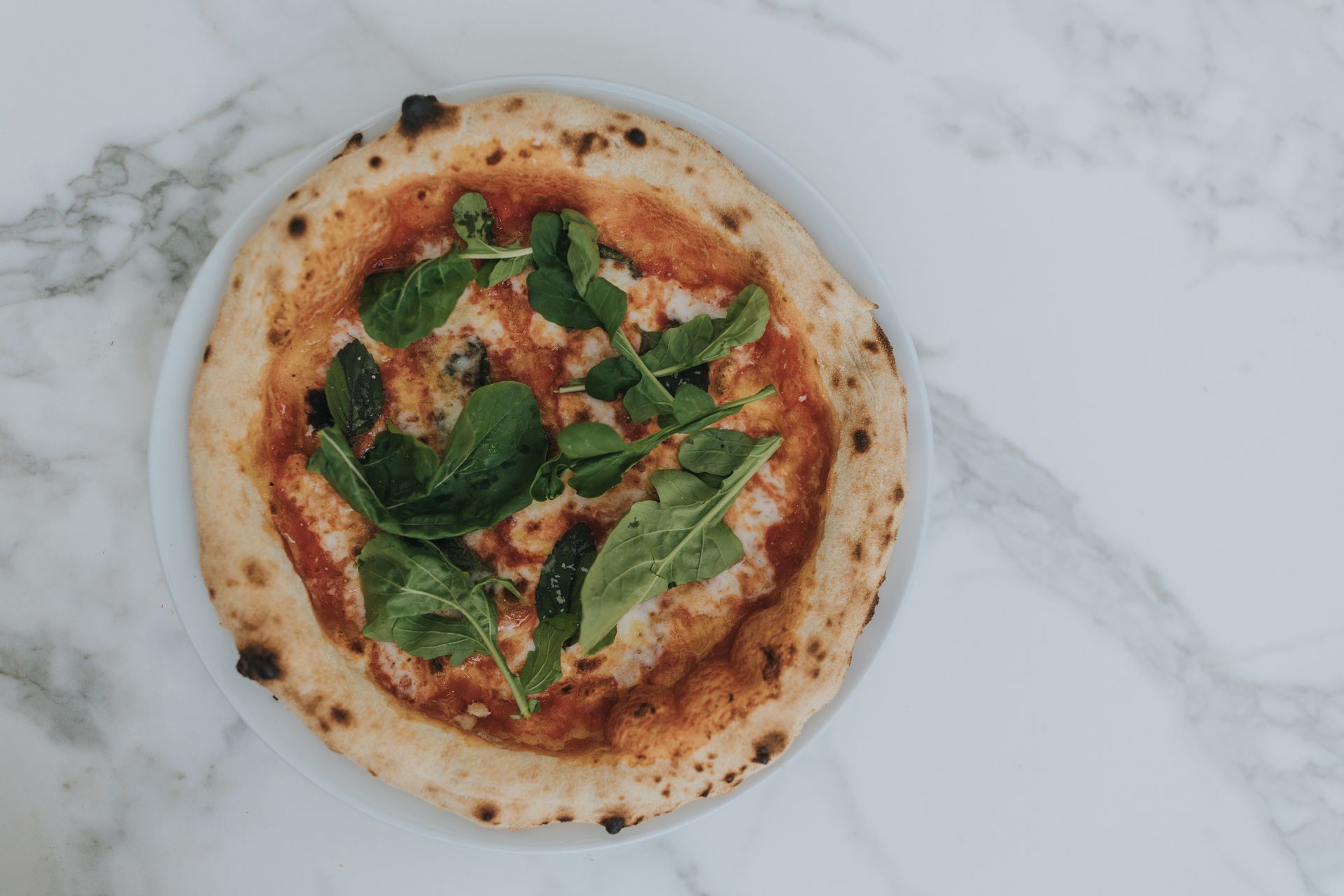 Pizza on a white plate, topped with tomato sauce and arugula, on a marble surface.